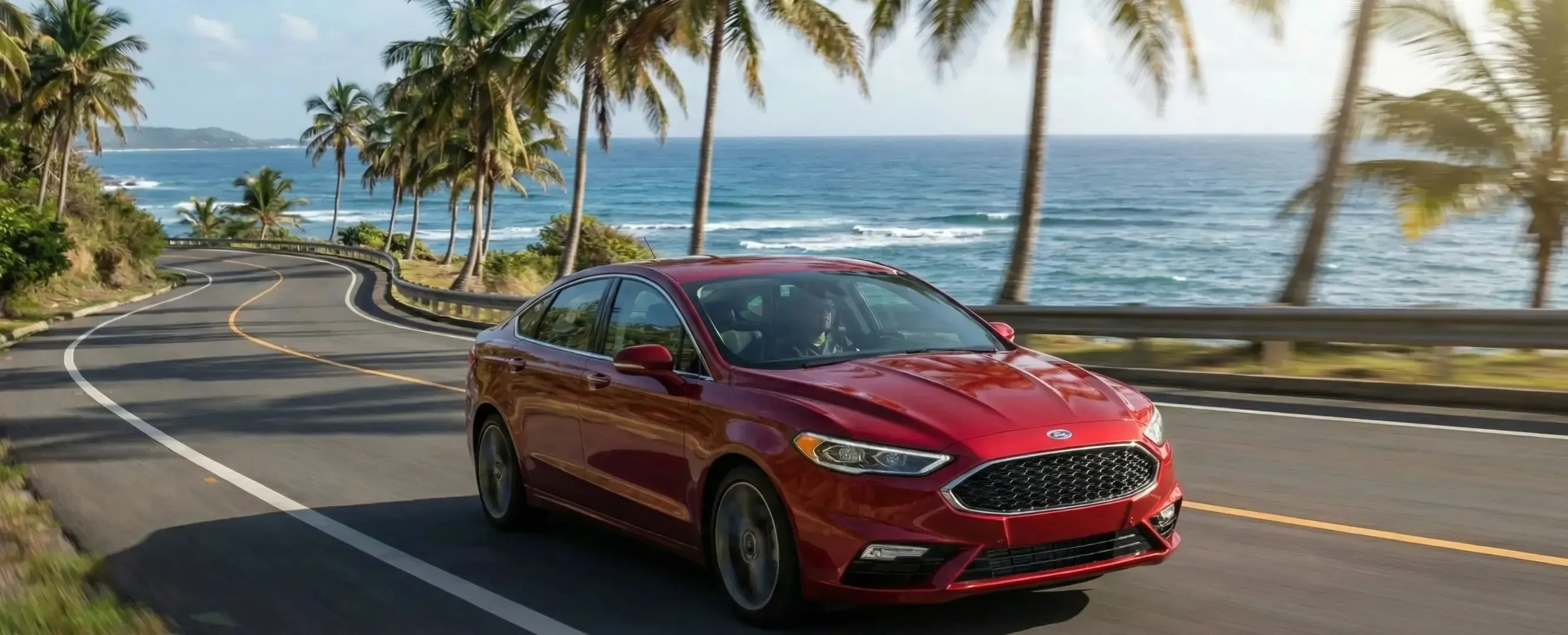 Red sedan driving a coastal California highway lined with palm trees